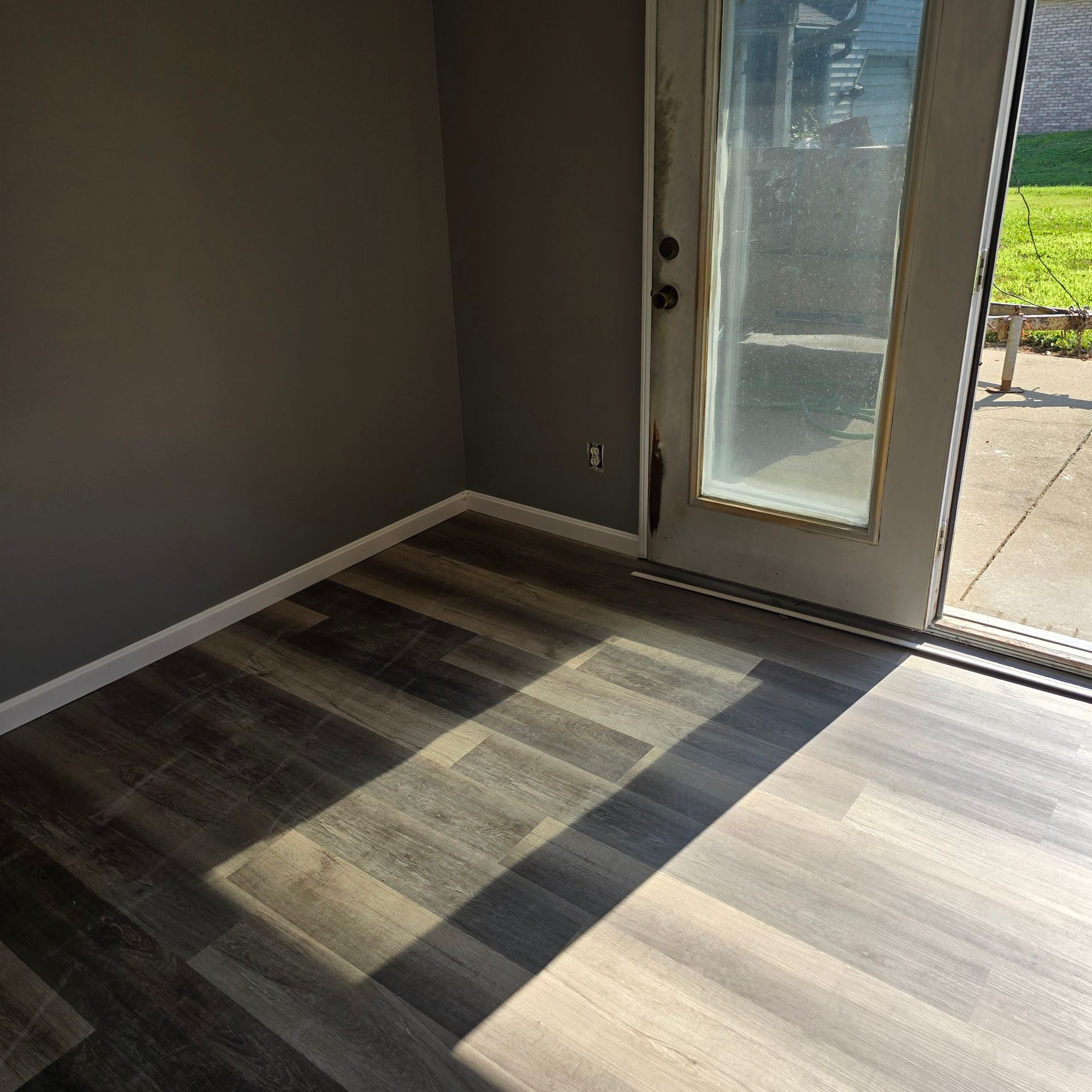 Dark gray room with wooden floor, sunlight, and a door leading outside.