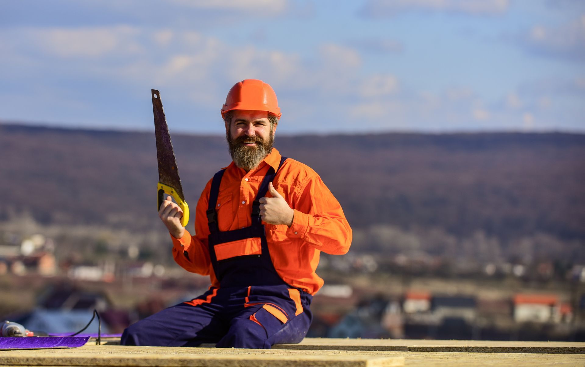 A man with a beard is kneeling down holding a saw and giving a thumbs up.