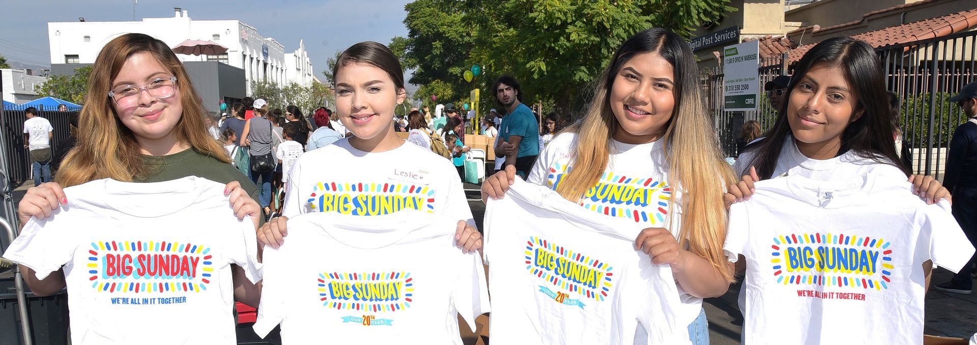 Four young people holding up white t-shirts with colorful text. Outdoors, likely at an event.