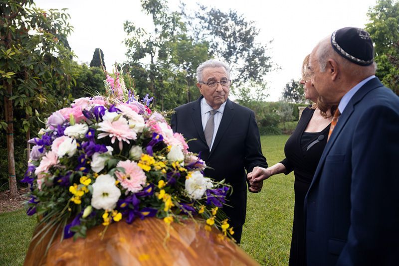 A man in a suit and tie is standing next to a woman in a black dress. looking at coffin with flowers