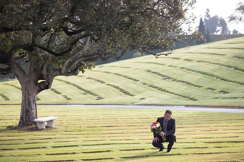 A man in a suit is kneeling in a cemetery next to a tree.