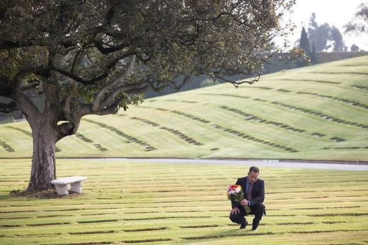 A man in a suit is kneeling in a cemetery next to a tree.