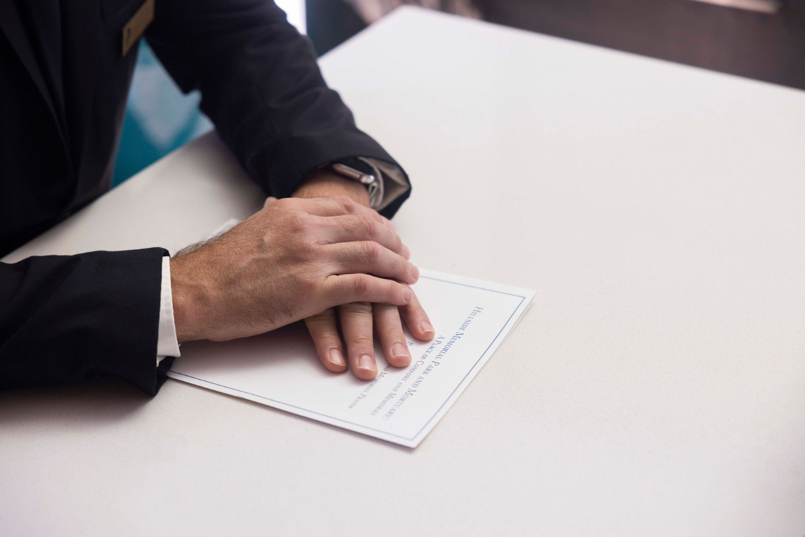 A man in a suit is sitting at a table with his hands folded on a piece of paper.