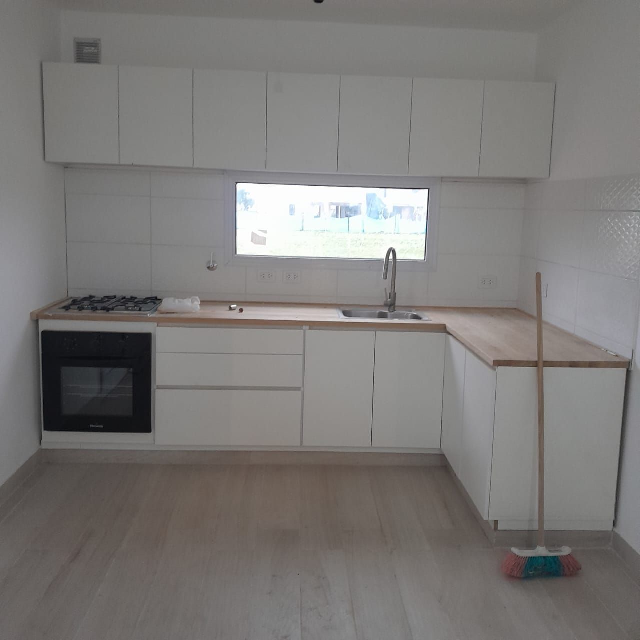 Cocina blanca en forma de L con encimeras de madera clara, ventana, horno y escoba en el suelo.