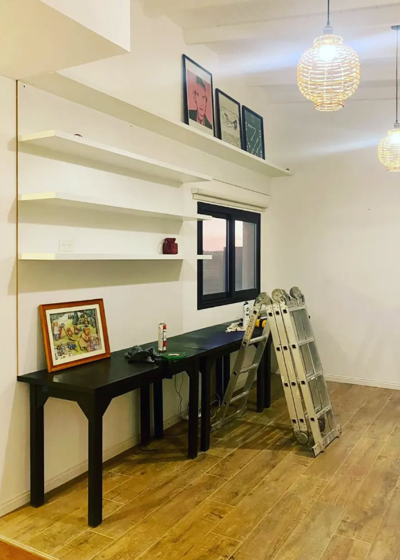 Habitación con estantes blancos, escritorio negro, ventana y escalera. Dos lámparas esféricas colgantes y cuadros en los estantes.