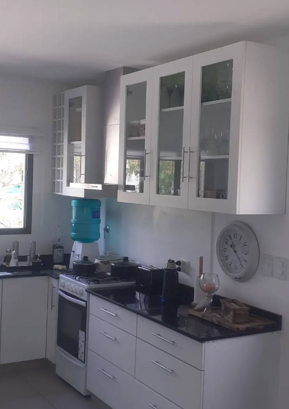 Cocina blanca con gabinetes altos, estufa y dispensador de agua. Encimeras negras y reloj de pared.