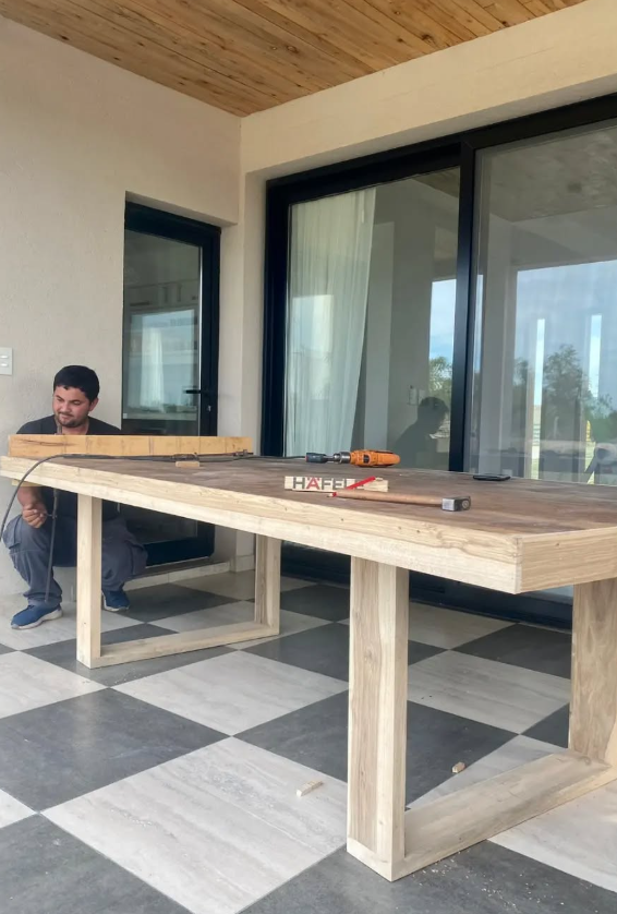 Un hombre trabaja en una gran mesa de madera en un patio. Sostiene una herramienta. La mesa tiene un suelo gris a cuadros.