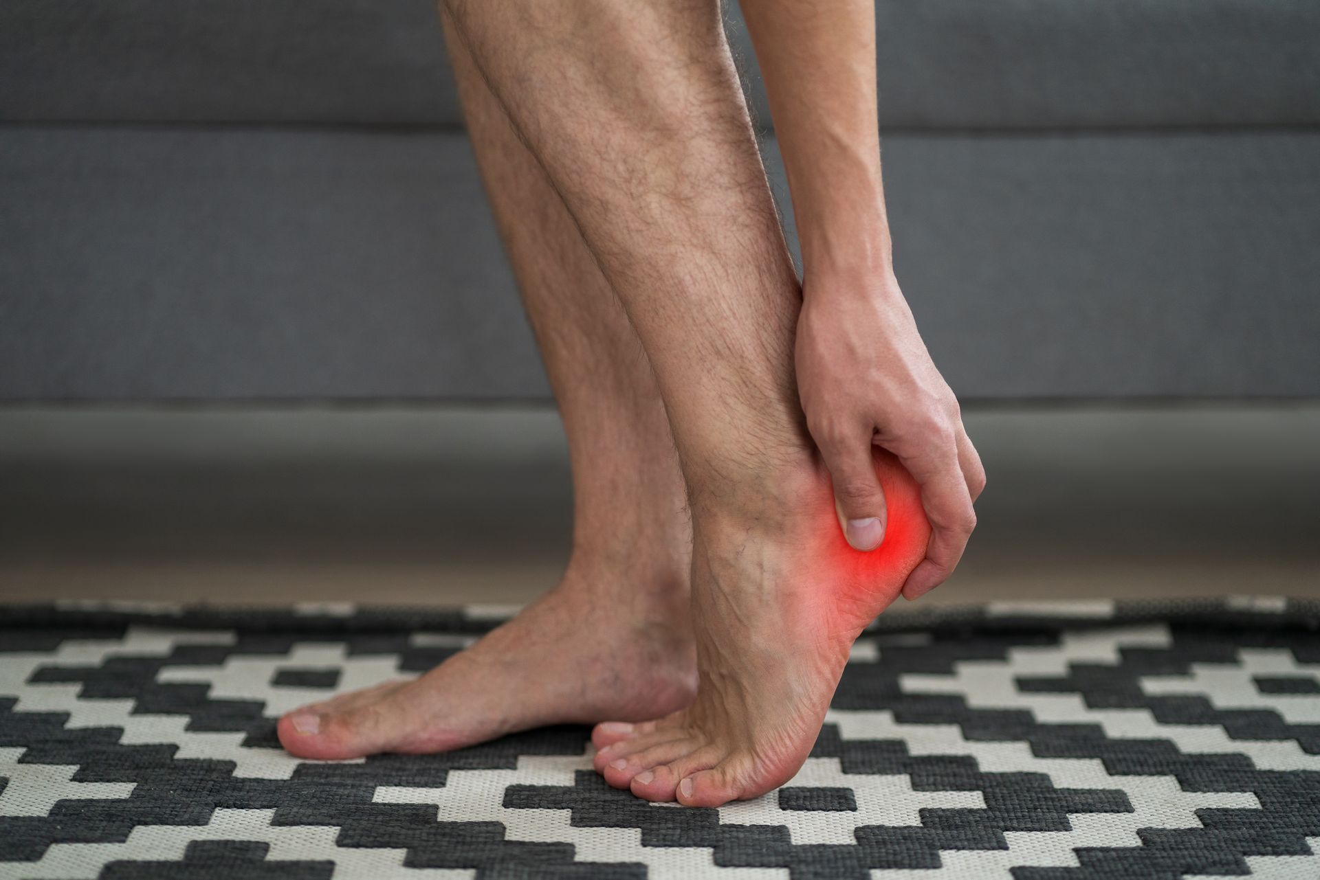 Man holding his heel, indicating pain, with a red highlight; indoors on a patterned rug.