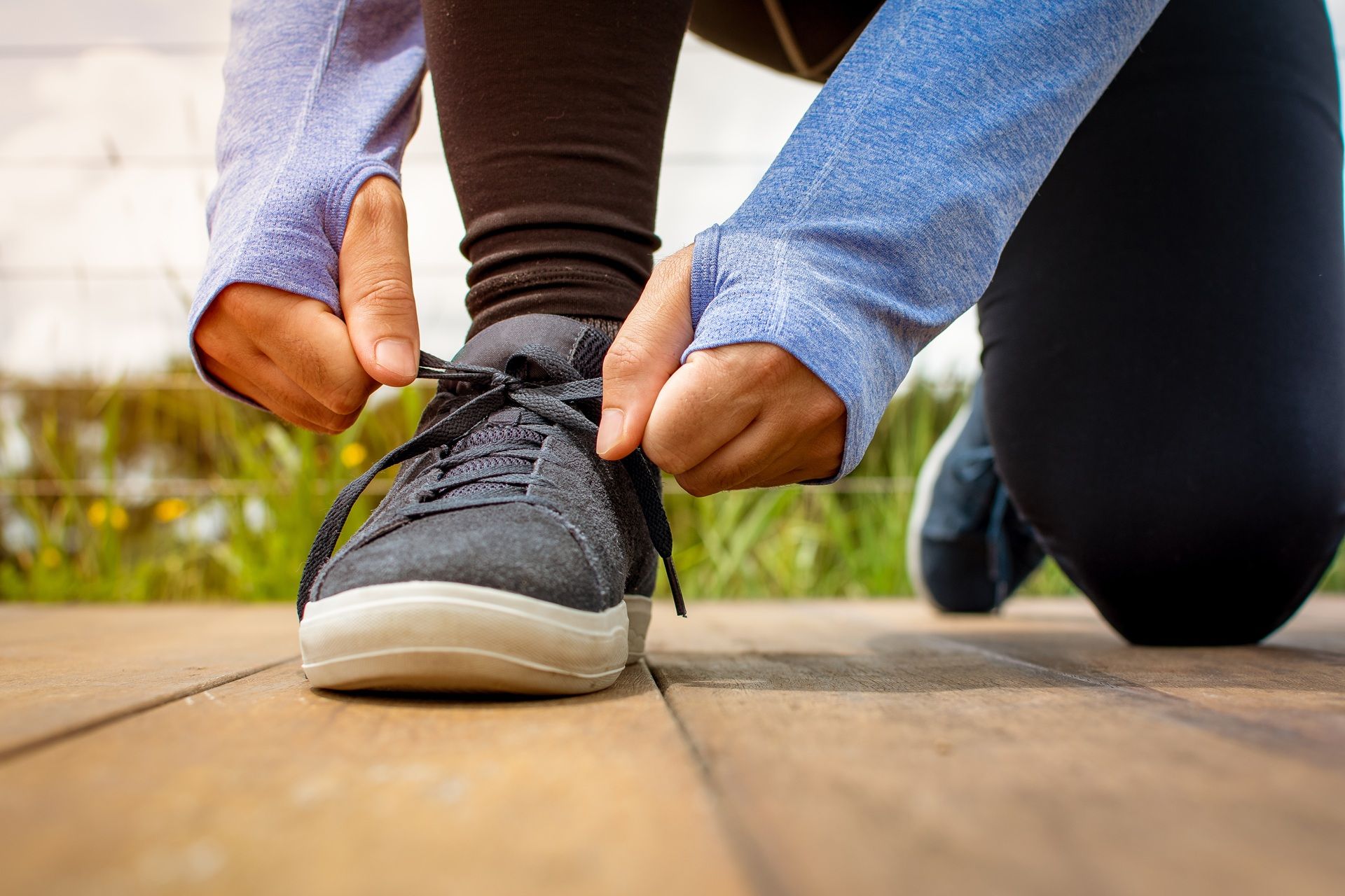Person kneeling on a wooden deck, tying the laces of a gray running shoe.