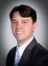 Man in a suit smiling, looking toward the viewer. He has dark hair and a blue tie.