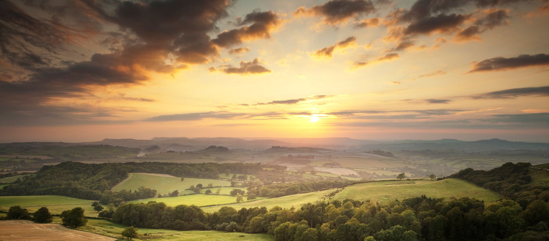 Sunset over a rolling green landscape with trees, fields, and hazy mountains; orange and yellow sky.