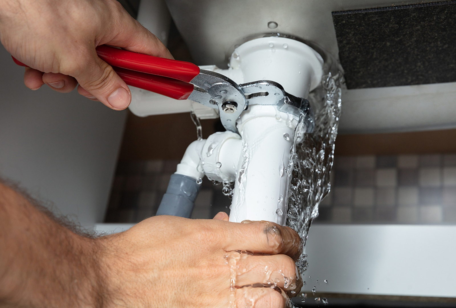 Hands using red wrench on leaking white plumbing under a sink.