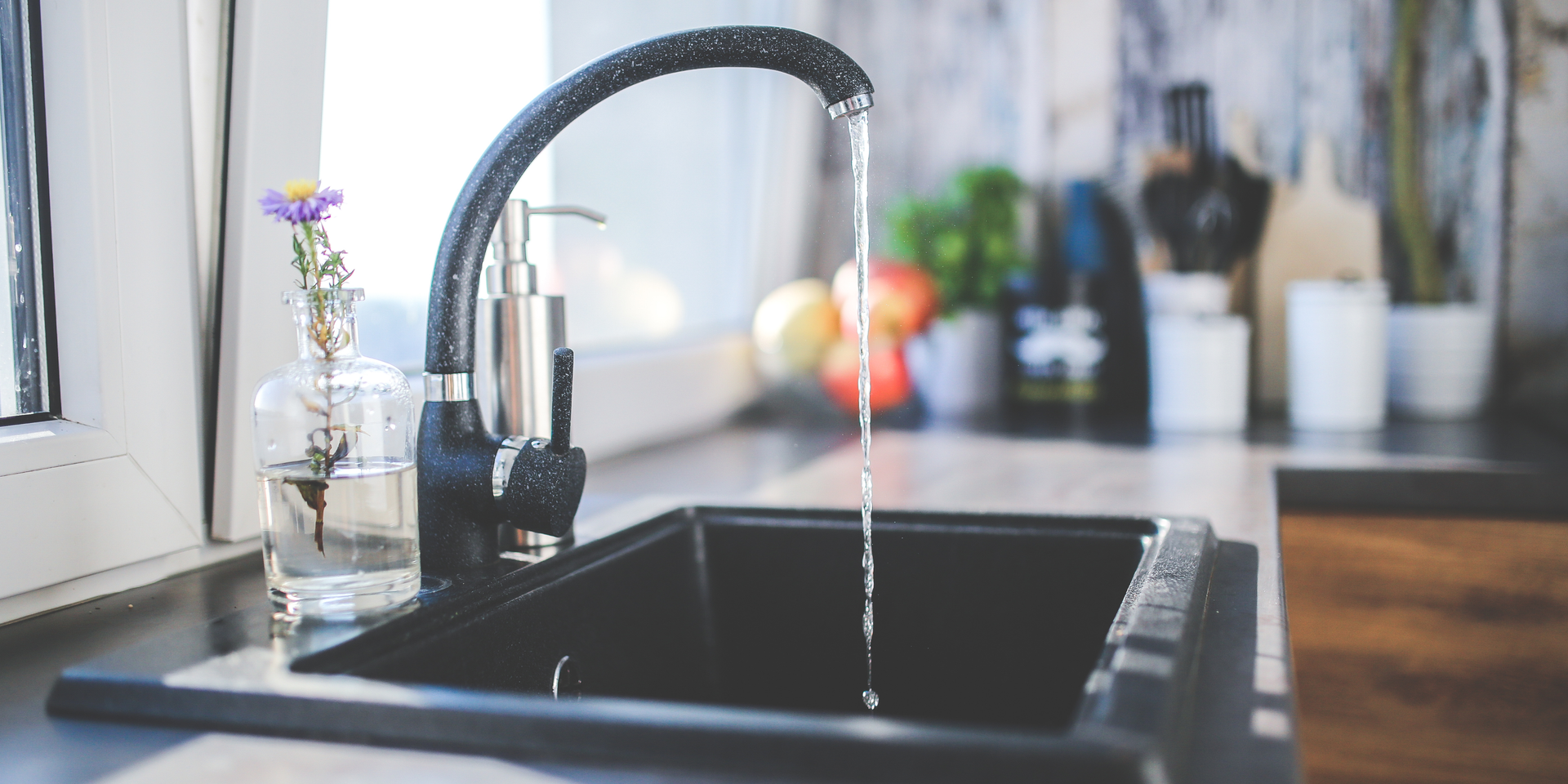 Black kitchen sink with running water from a black faucet, beside a vase with a flower.