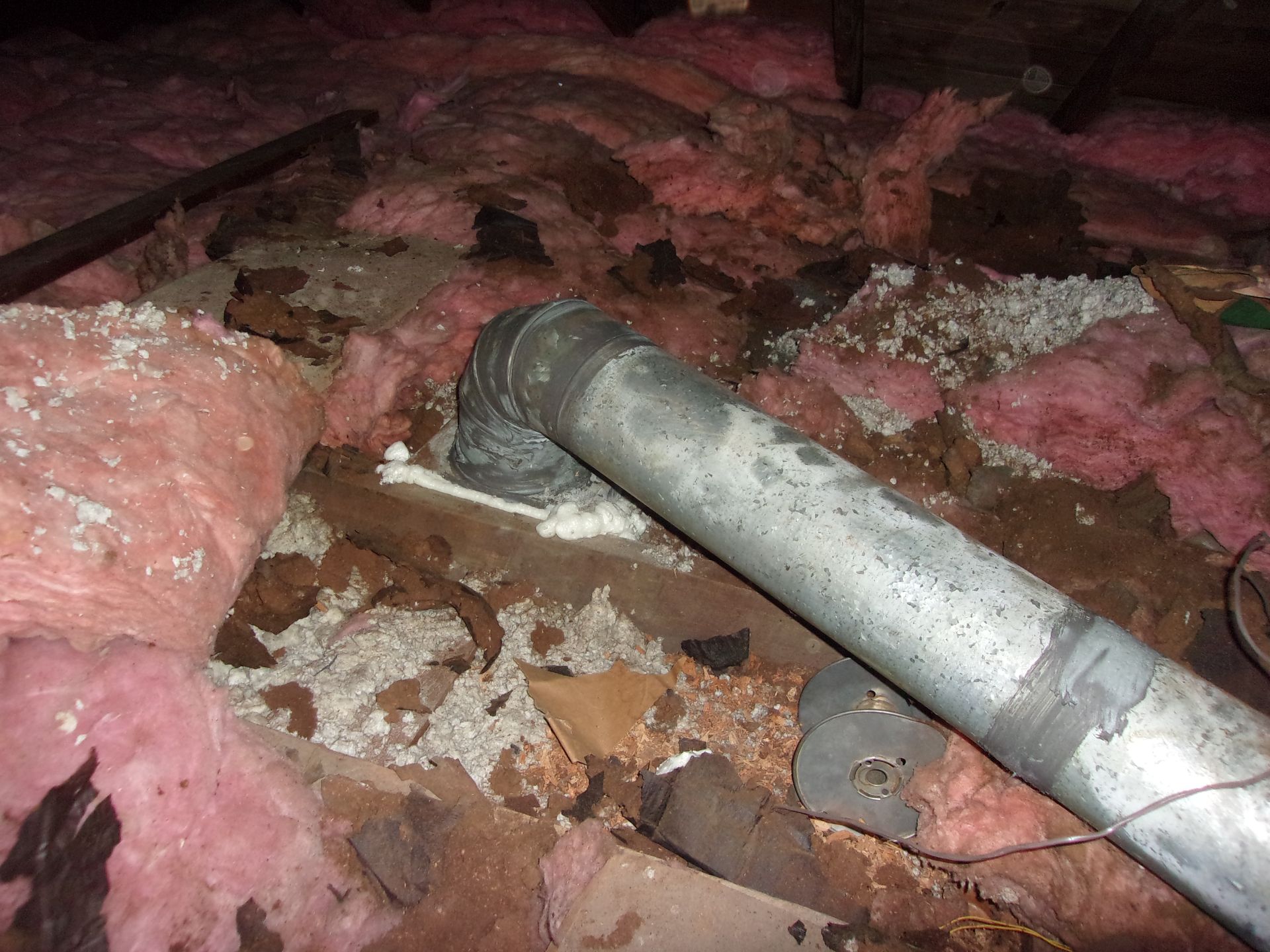 Attic view with pink insulation, a metal vent pipe, and possible water damage visible on wood and insulation.