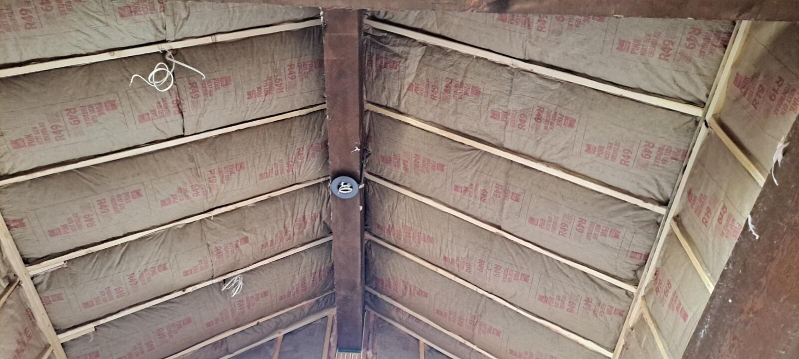 View of an attic ceiling with insulation between wooden beams. A light fixture is mounted on a central beam.