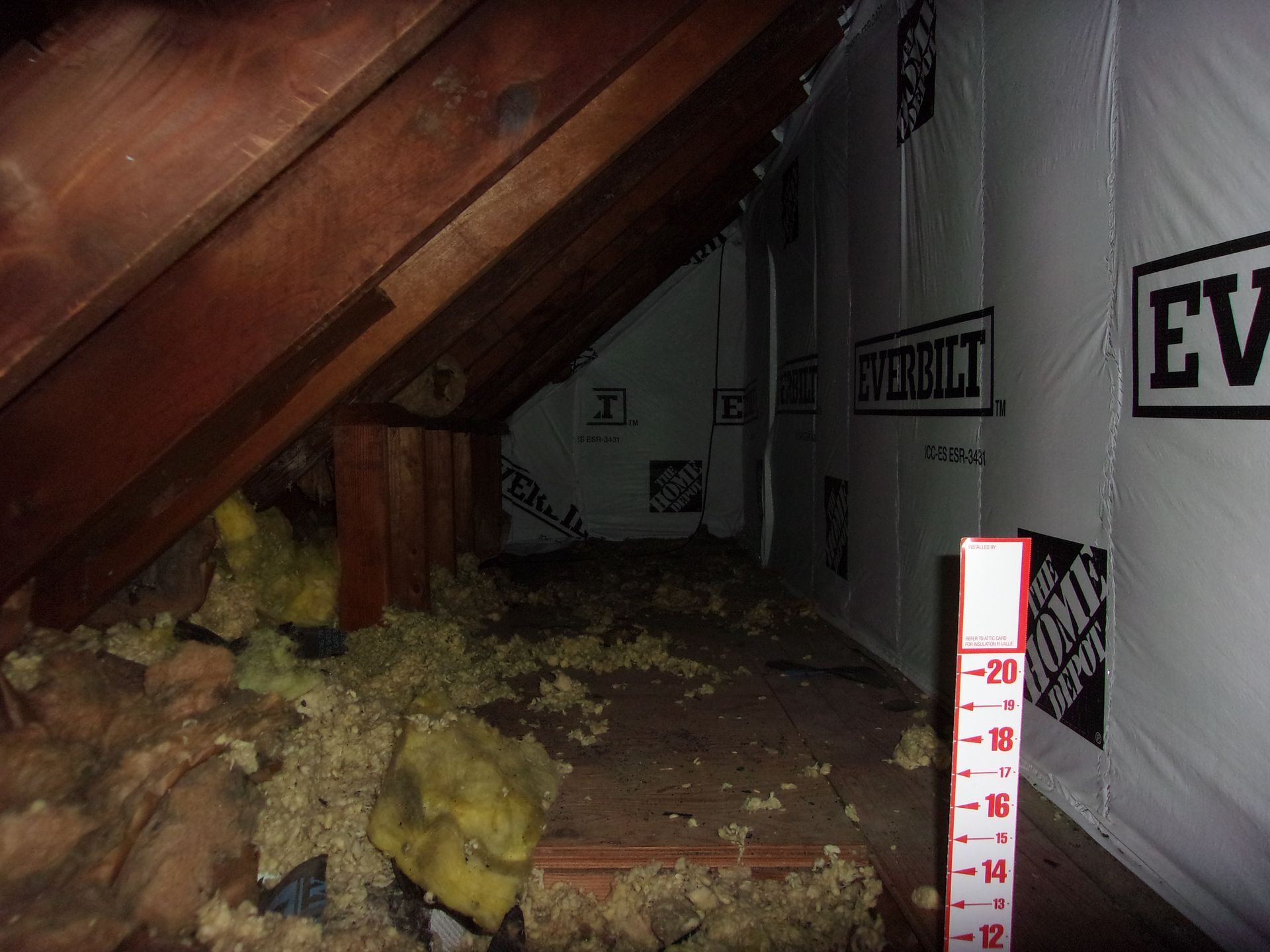 Interior view of an attic. Visible are wooden beams, insulation, and vapor barrier, with a Home Depot brand measuring stick.