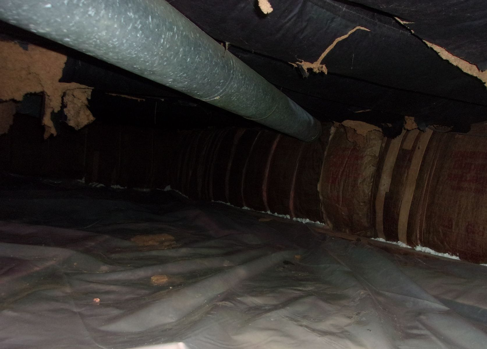 Dark crawlspace with plastic sheeting on the ground and insulated ductwork and pipes running along the ceiling.