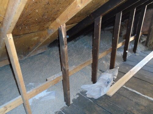 Attic with wooden beams, insulation, and a partially visible floor. The space is dimly lit.