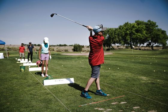 Boy in red shirt swings a golf club on a sunny driving range. Other golfers watch nearby.