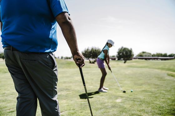 Man in blue shirt watches a young girl in purple shorts as she prepares to swing a golf club on a sunny green.