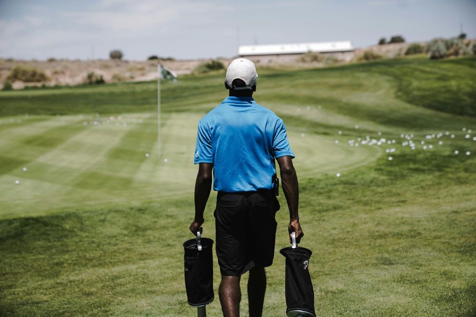 A person in a blue shirt and shorts walks on a green golf course, carrying two black ball holders.