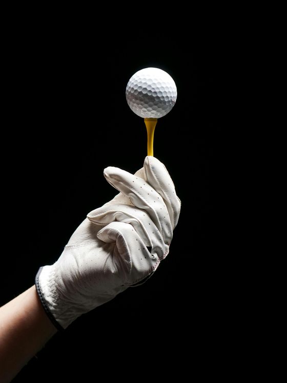 Hand wearing a golf glove holding a golf ball balanced on a tee against a black background.