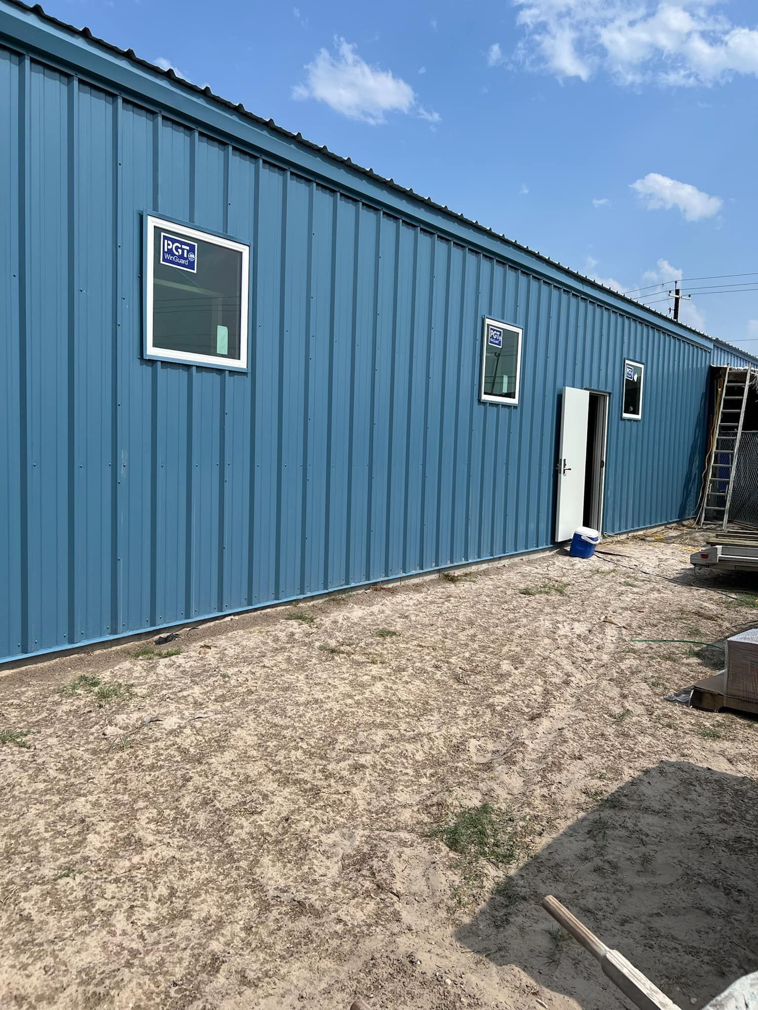 A long blue metal-sided building under a bright blue sky, with three windows, a door, and a ladder on a gravel lot.