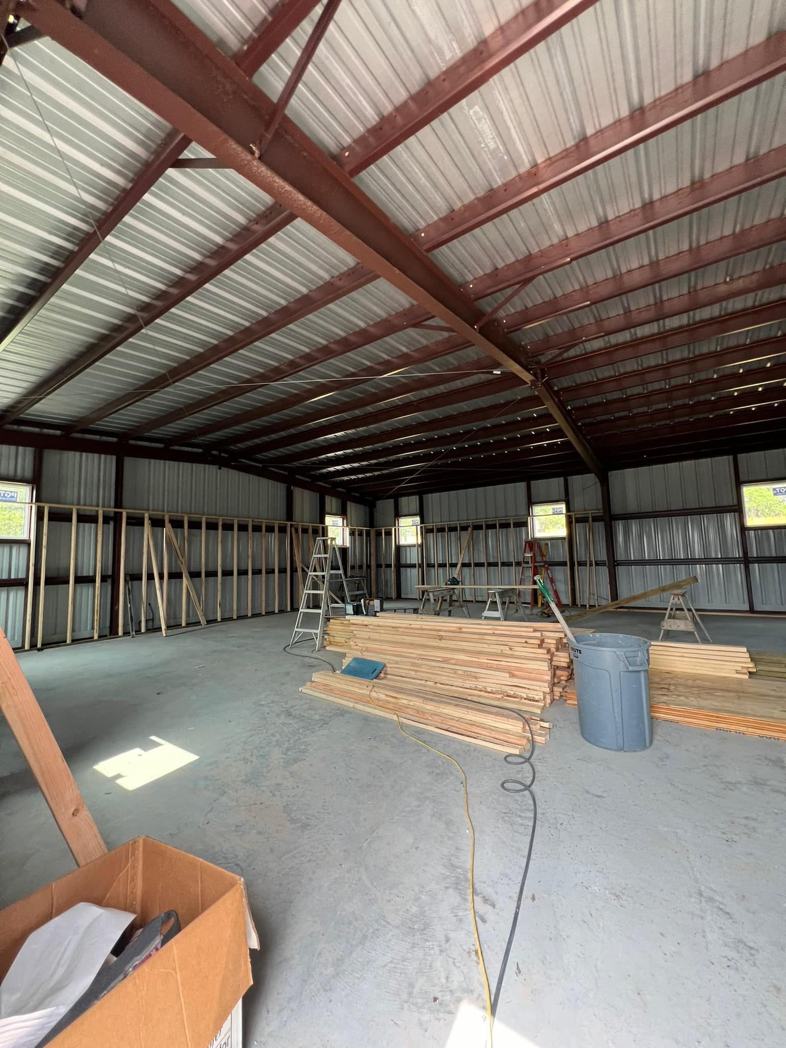 A large, unfinished metal building interior featuring exposed steel rafters, corrugated walls, and construction materials.