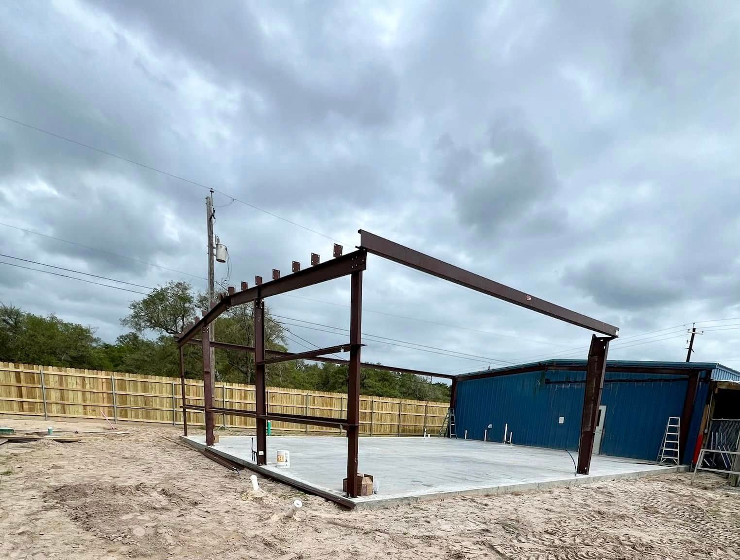 Steel framework of a building under construction on a concrete slab against a cloudy sky.
