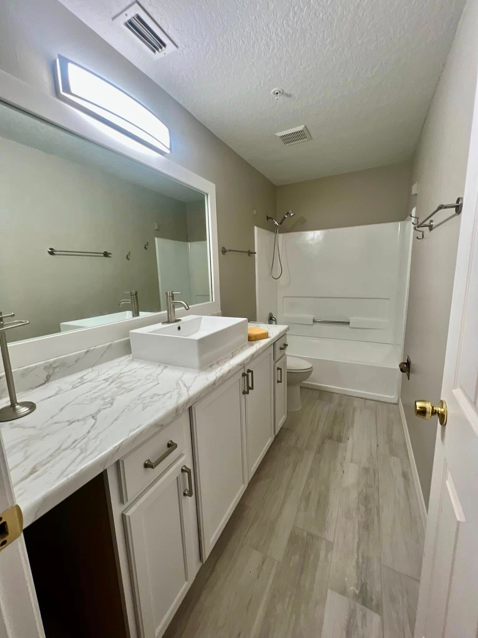 A bright bathroom with a white vessel sink on a marble-style counter, a large mirror, and a walk-in shower with light floors.