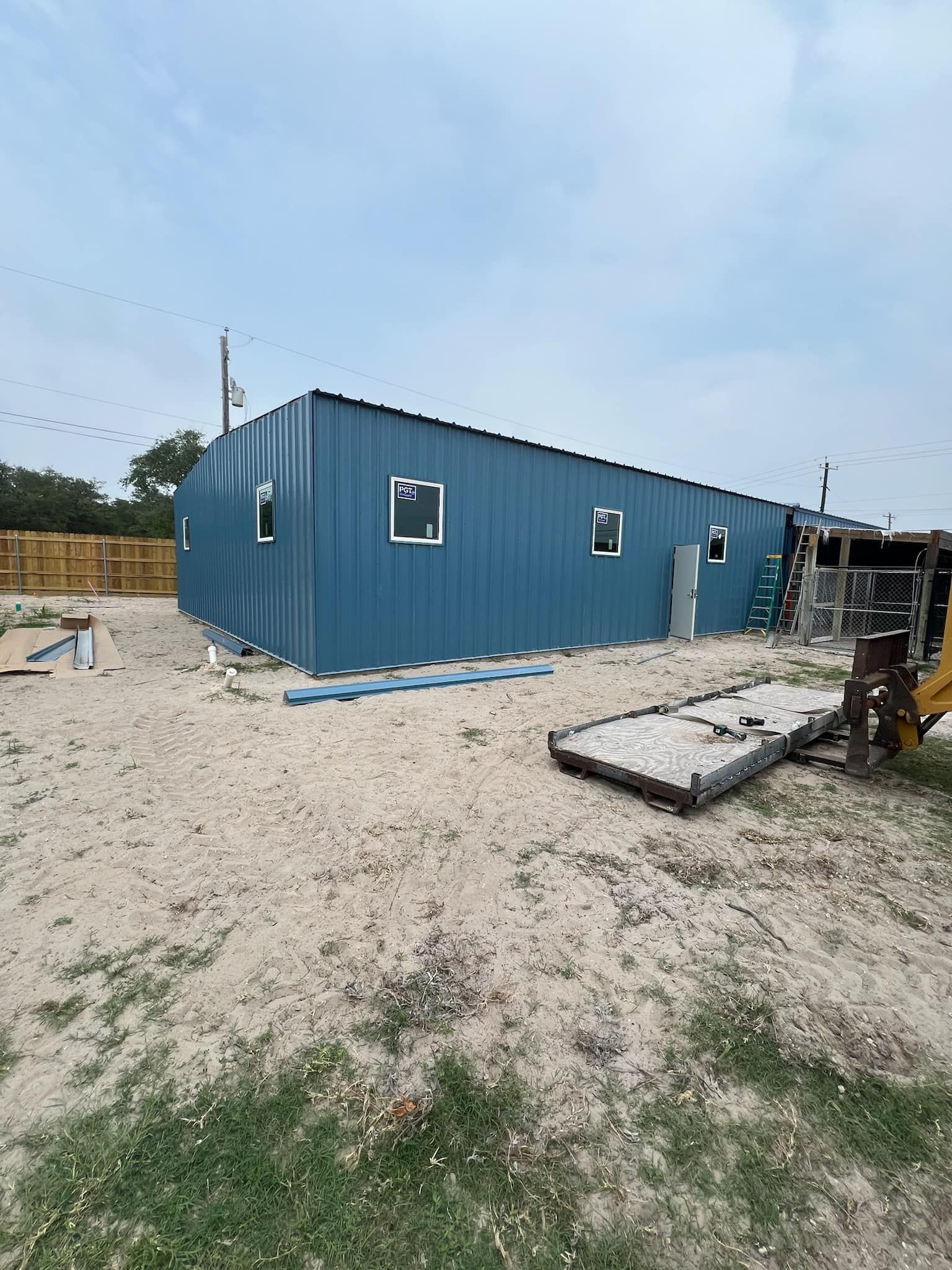 A long, blue metal-sided building with windows, under a blue sky on a sandy, partially grassy construction site.