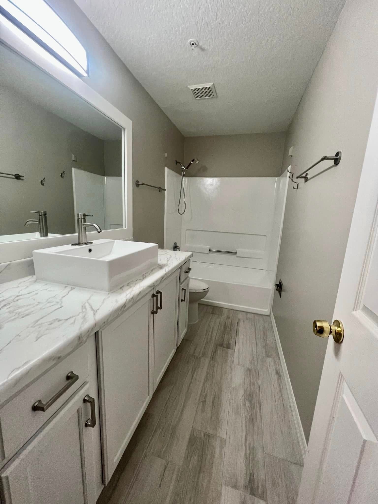 A bright bathroom with a white marble-top vanity, vessel sink, rectangular mirror, and a white shower-tub combination.