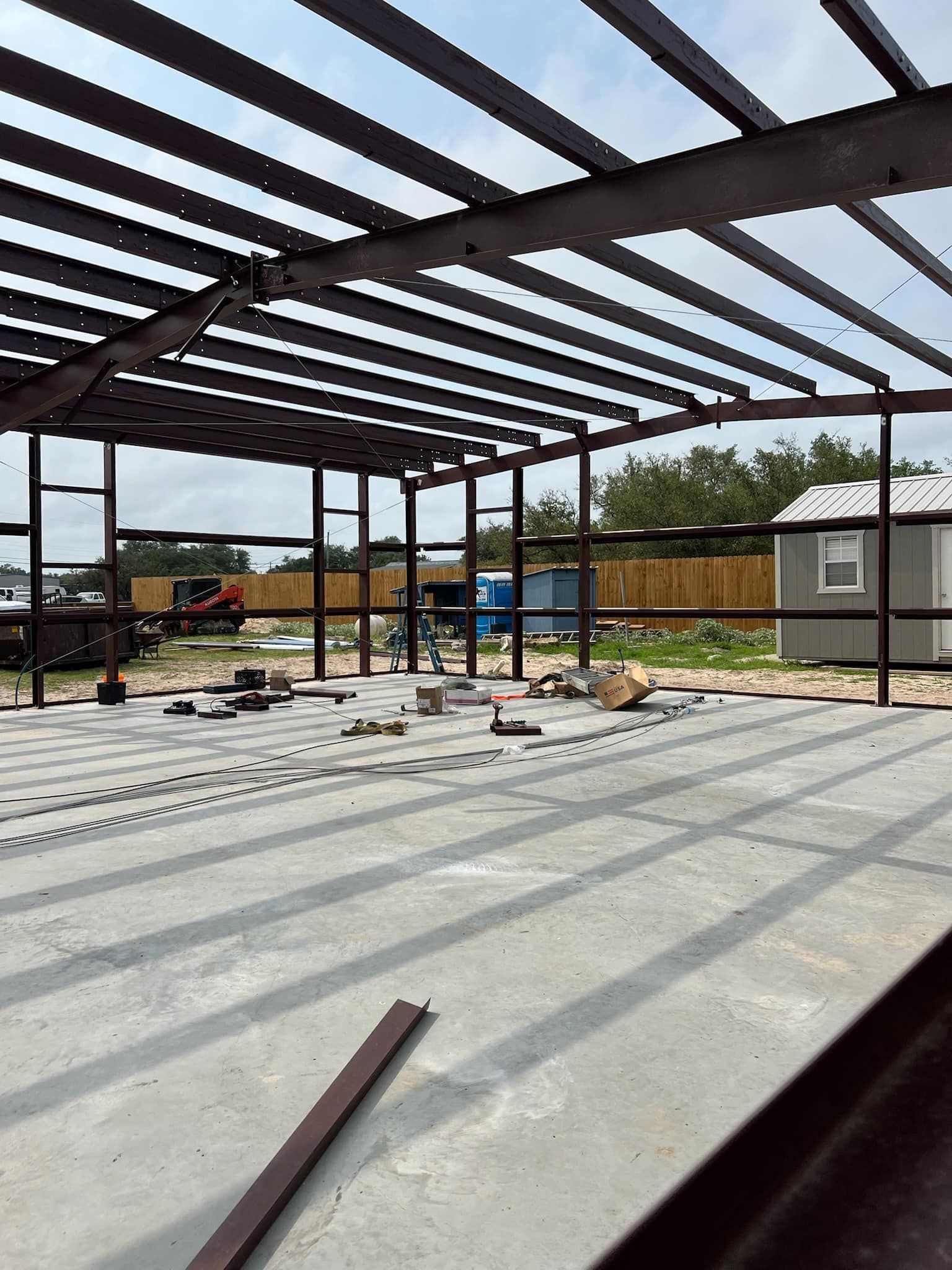 A large, open-air metal building under construction, featuring exposed steel beams and a level concrete floor.