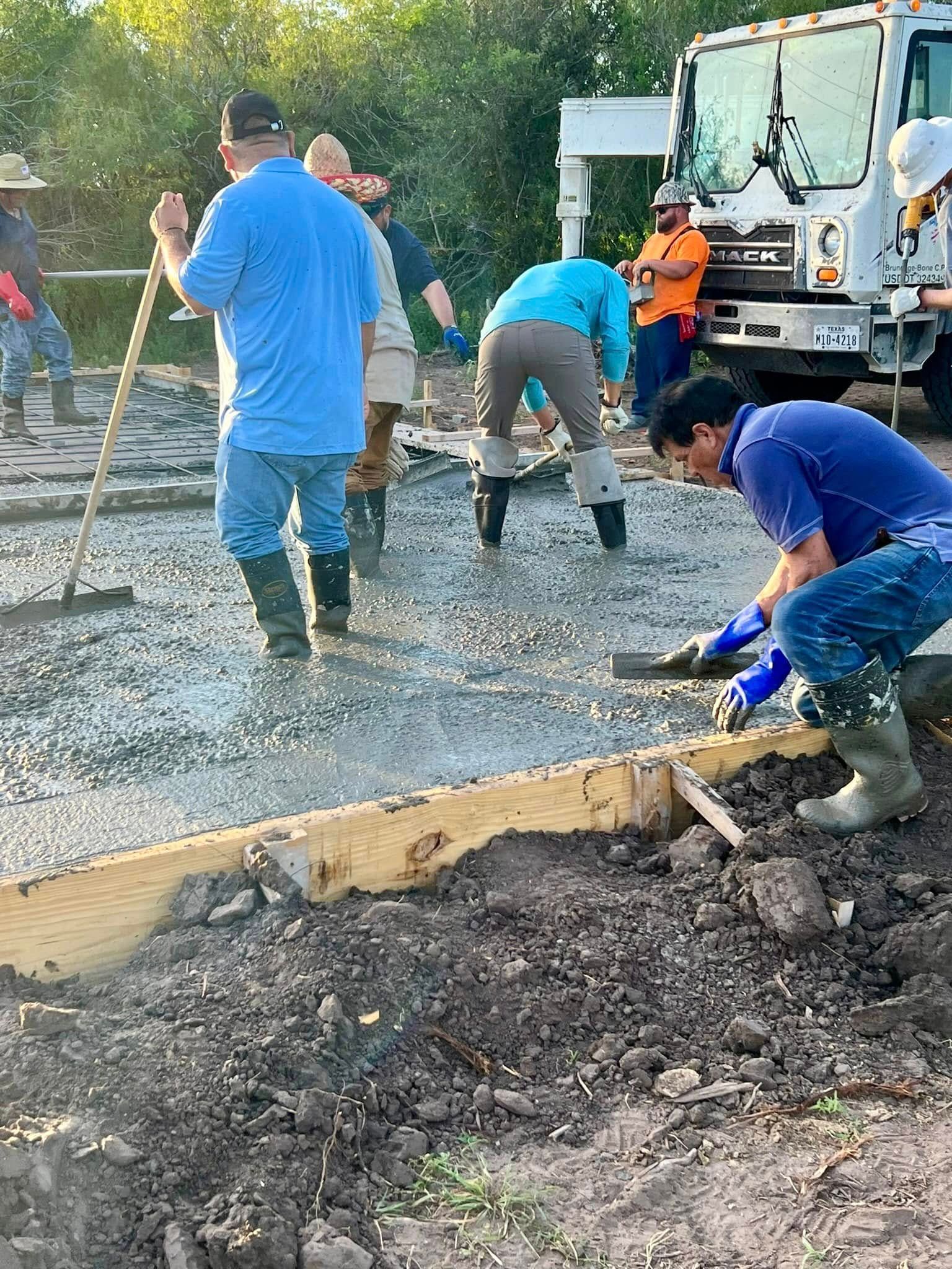 A group of people wearing work clothes and boots smooth wet concrete inside wooden forms near a cement mixer truck.