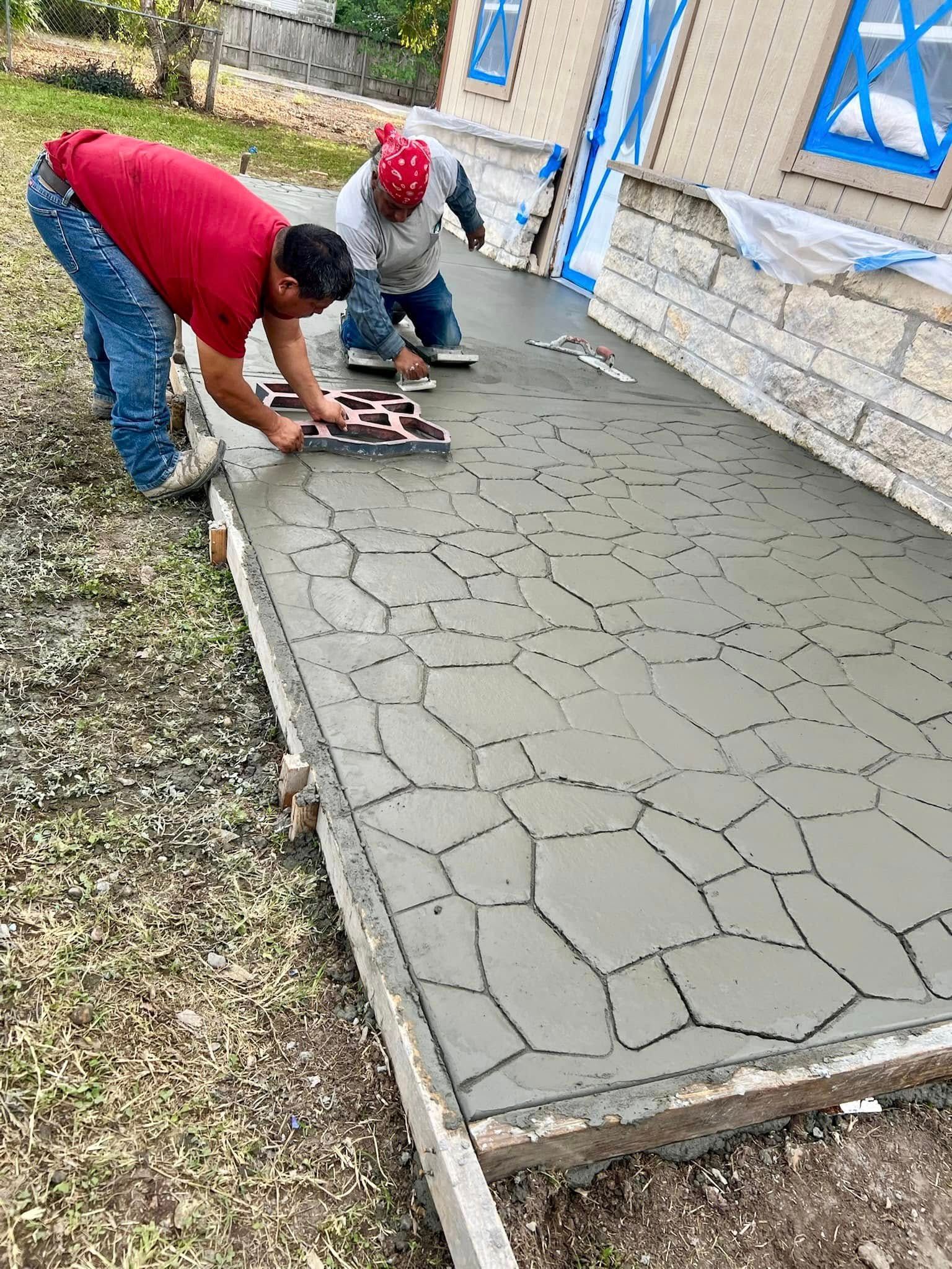Two workers use a patterned stamp tool to imprint a decorative stone design into freshly poured wet concrete.