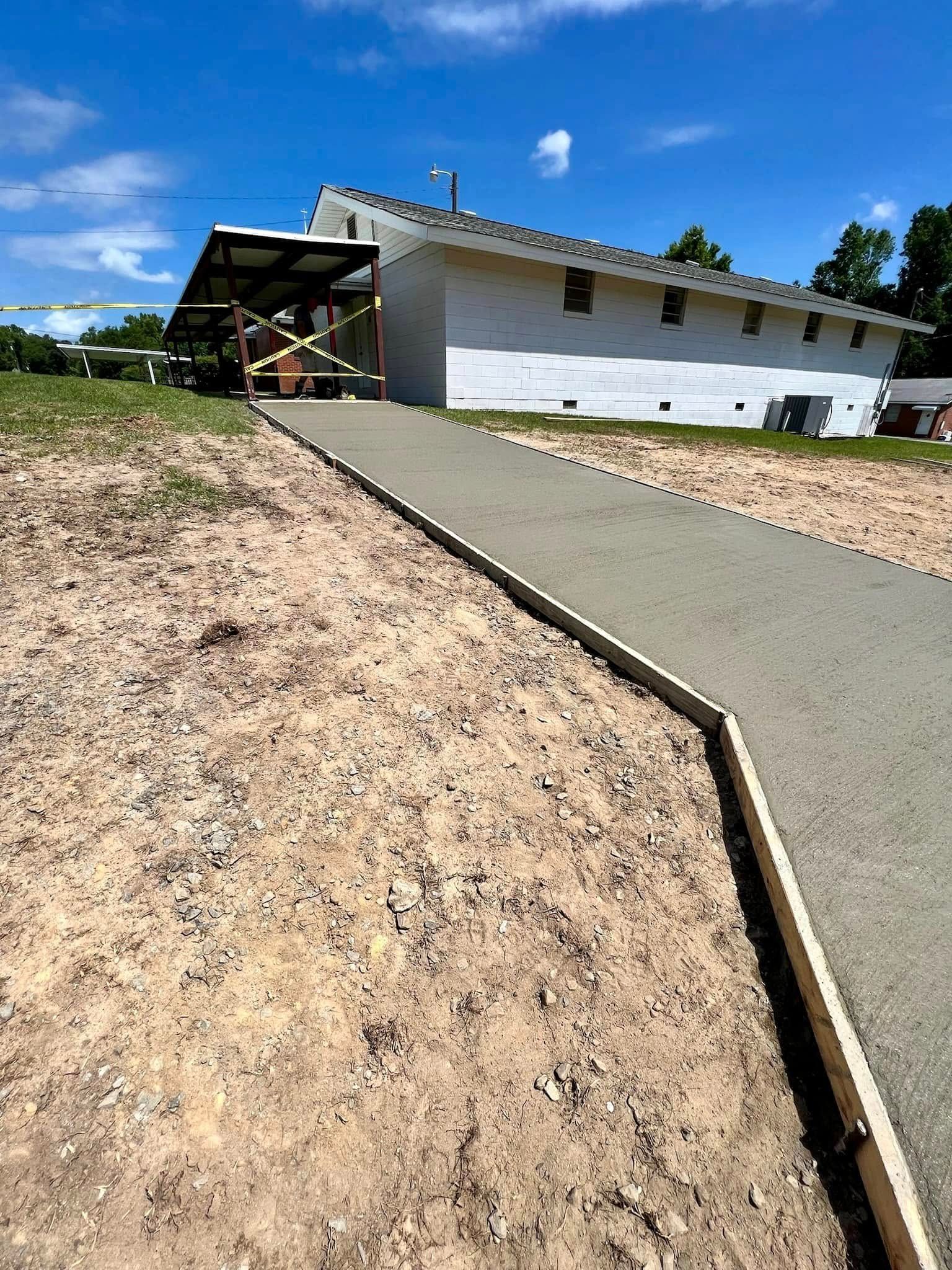 A newly poured concrete walkway leads toward a white, single-story building with a covered entrance under a blue sky.