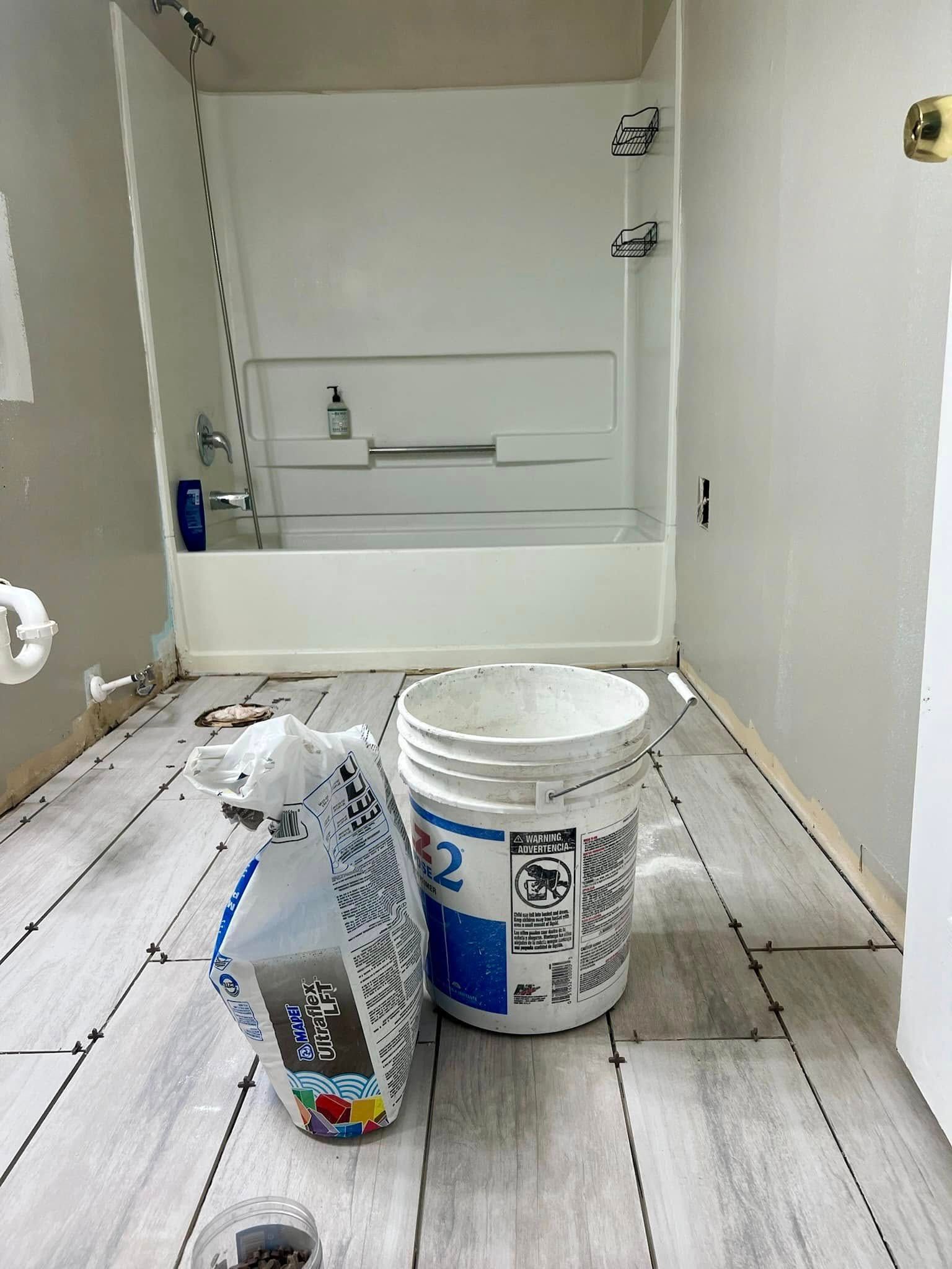 A bathroom with a white tub-shower unit, partially installed gray wood-look floor tiles, a bucket, and a bag of supplies.