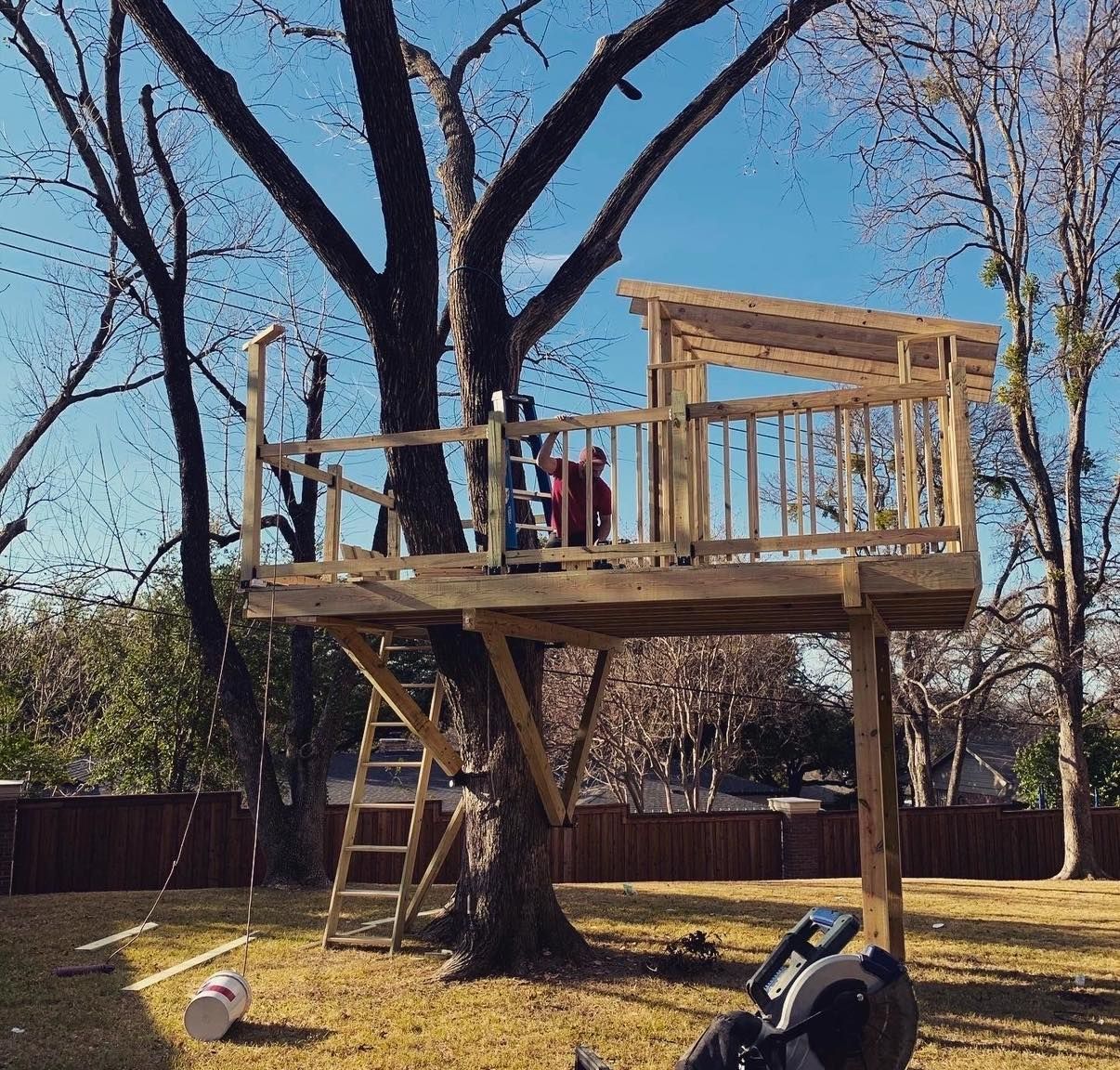 A person works on a wooden treehouse platform under construction in a large backyard on a sunny day.