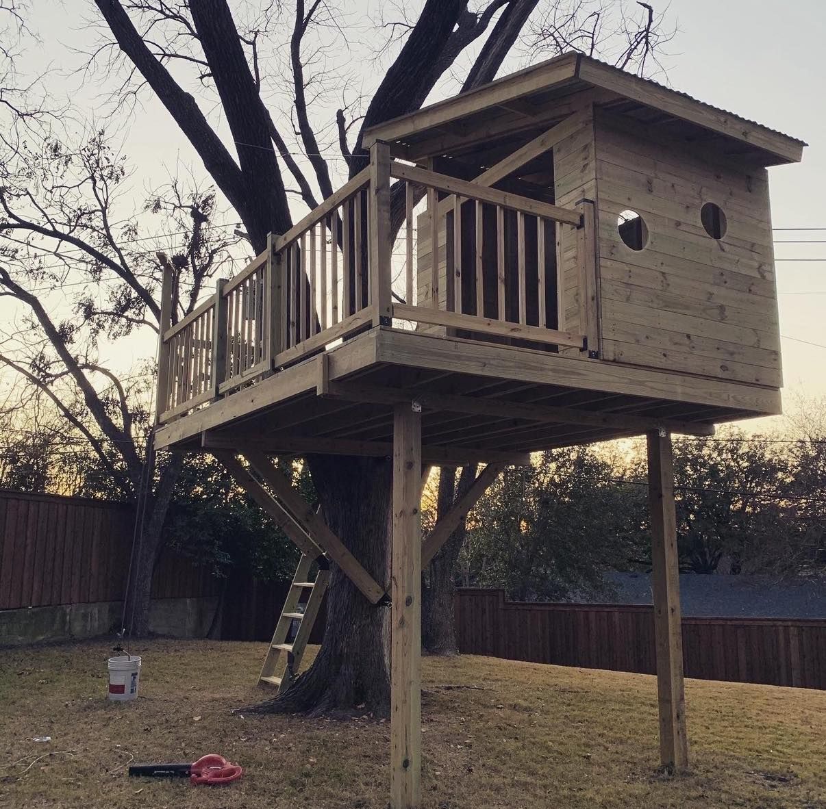 A wooden treehouse with a deck, railing, and roof, supported by a large tree and two vertical posts in a grassy yard.