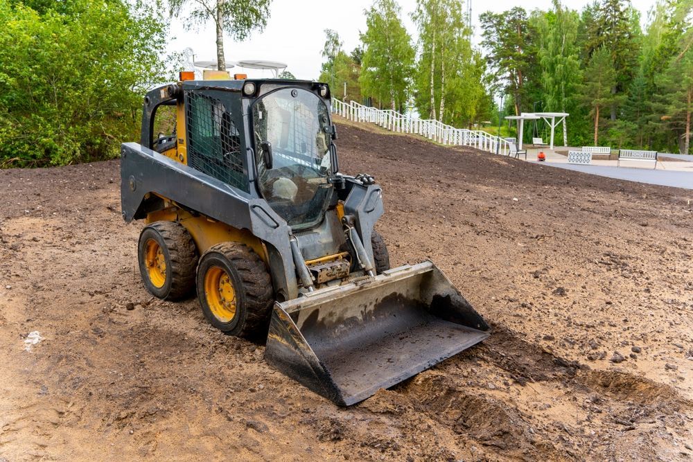 A yellow and gray skid steer loader with its bucket down on a muddy construction site near a forest and white fence.