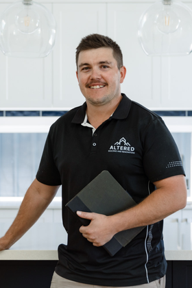 A person wearing a work shirt, cap, and tool belt stands with hands on hips inside a wooden construction frame.