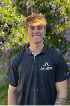 A person in a black t-shirt and work belt stands at a construction site with exposed wooden framing in the background.