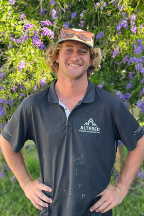 A person in a black t-shirt and work belt stands at a construction site with exposed wooden framing in the background.