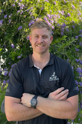 A smiling person with a red beard wearing a black polo shirt and tool belt, standing in a partially built wooden frame.