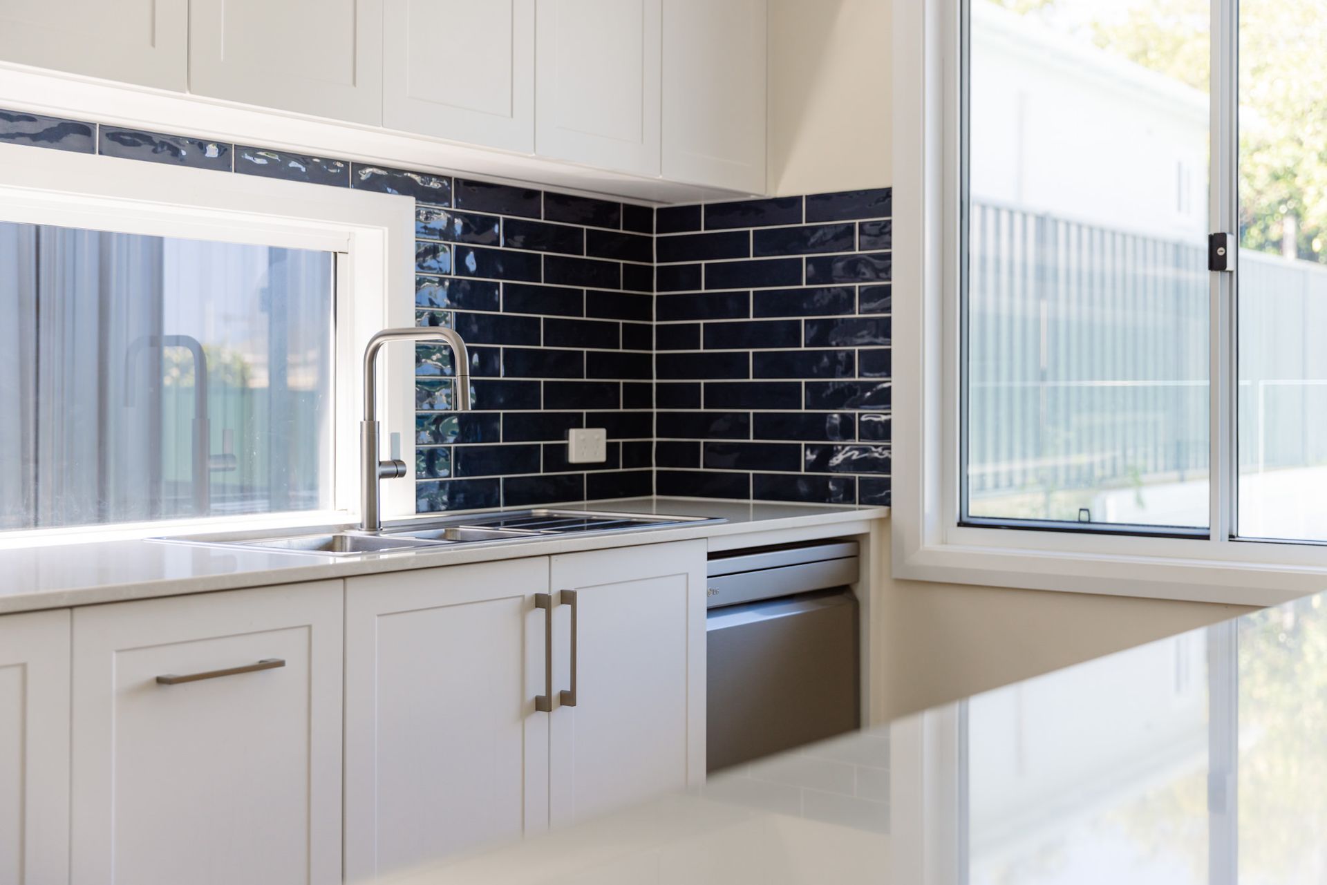 A modern white house with wooden accents and a gabled roof, set back from a paved driveway surrounded by green trees.
