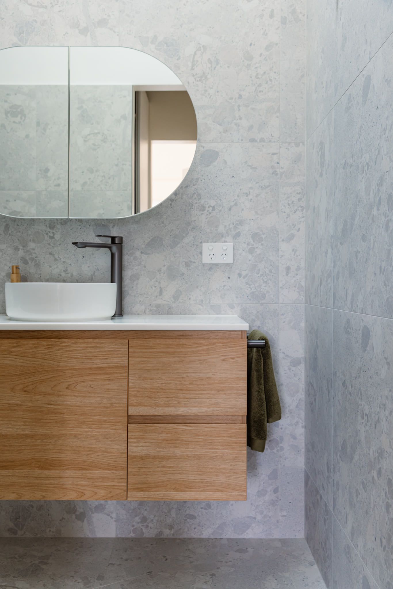 A modern bathroom vanity with a white vessel sink, dark metal faucet, and soap dispenser against gray terrazzo-style tile.