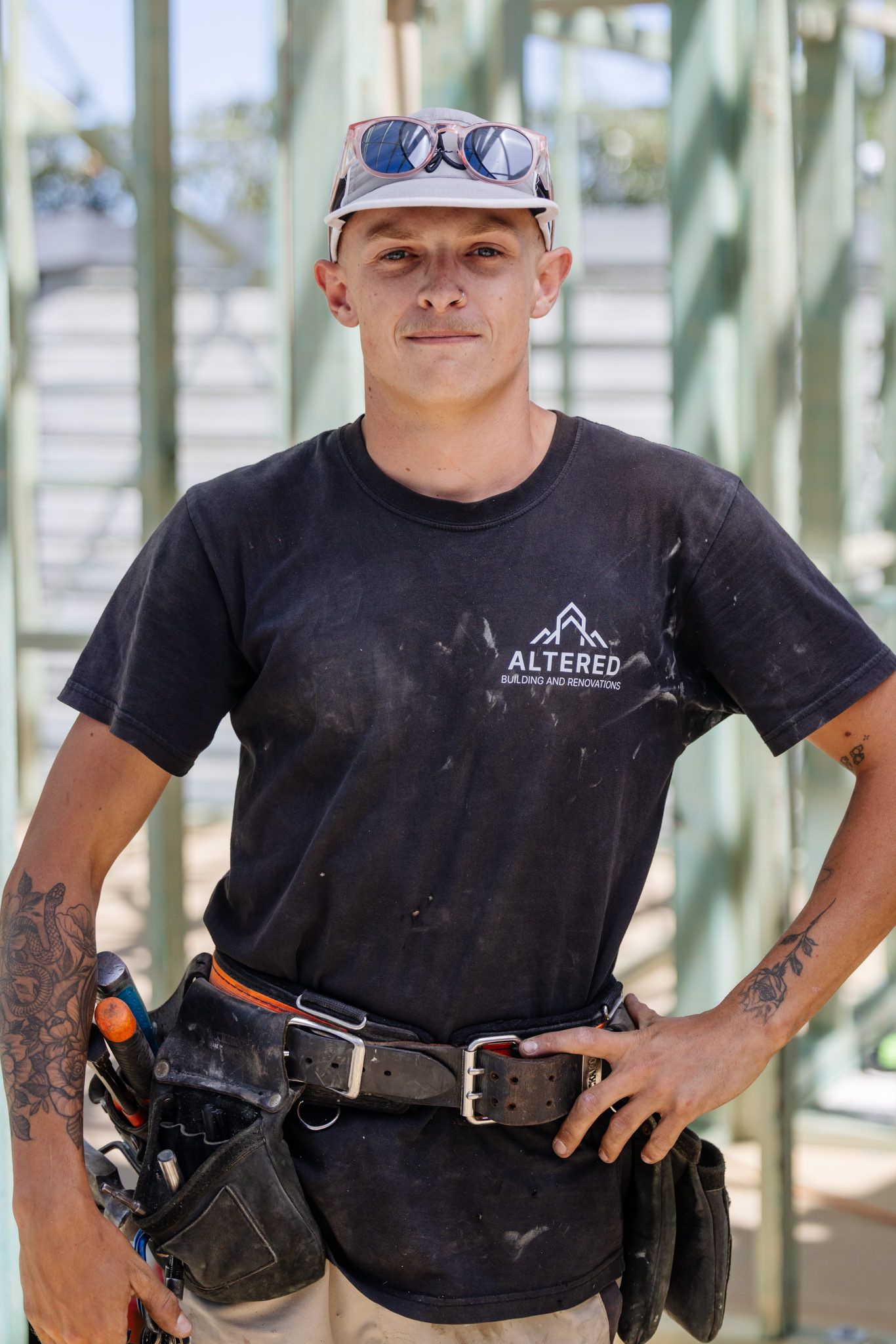 A person in a black t-shirt and work belt stands at a construction site with exposed wooden framing in the background.