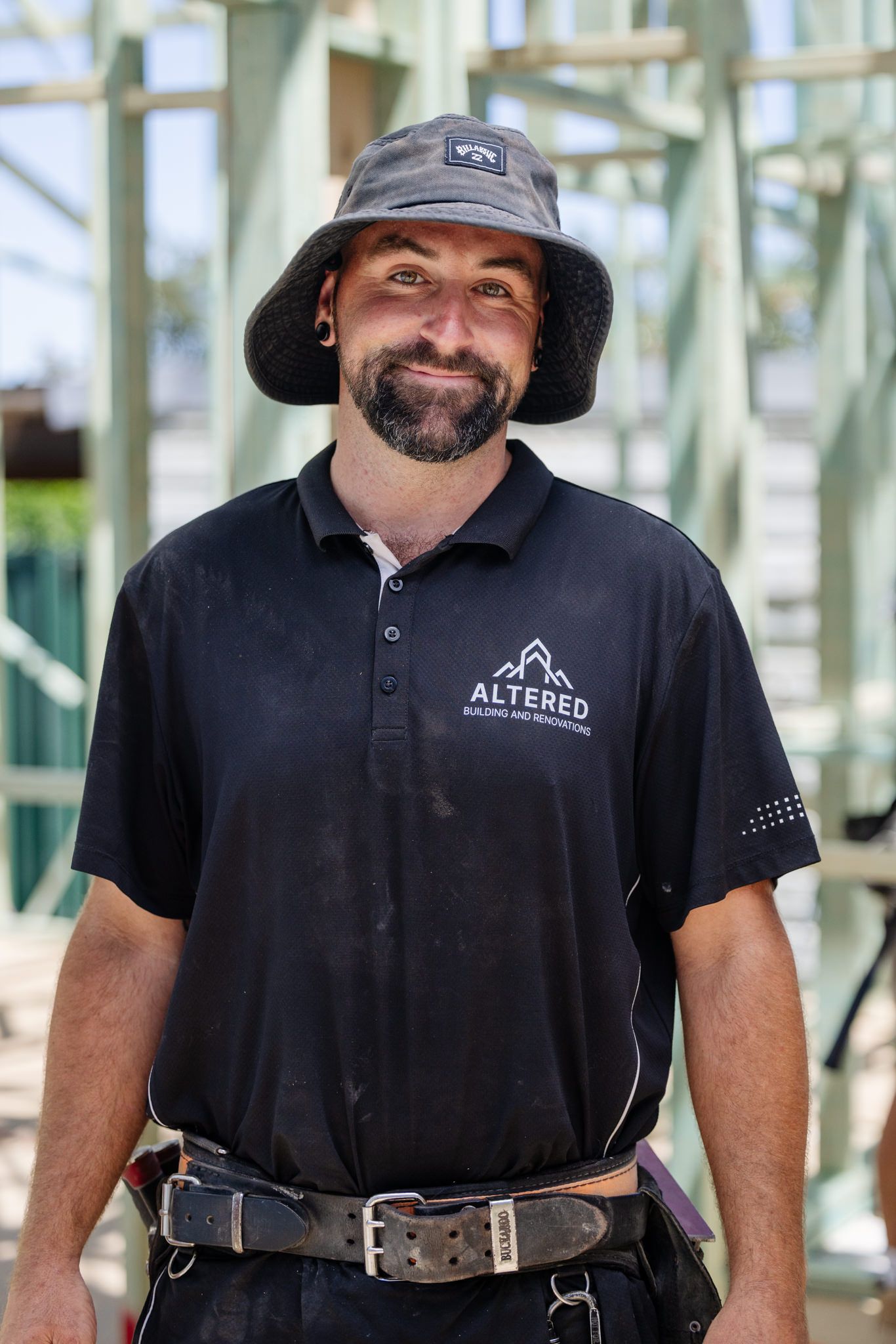 A person in a black polo shirt and bucket hat standing in front of a wooden building frame.