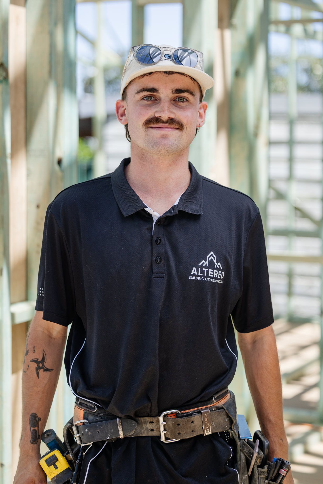 A smiling construction worker wearing a black polo shirt, tool belt, and cap, standing in a partially framed building.