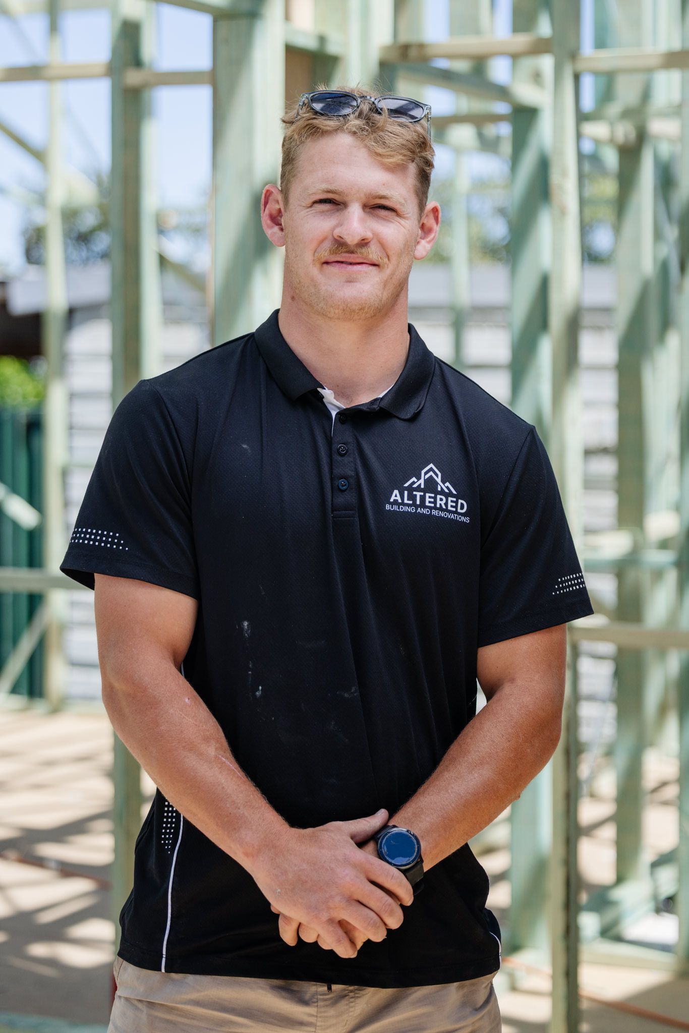 A person wearing a black polo shirt stands in front of a wooden building frame, hands clasped, looking at the camera.